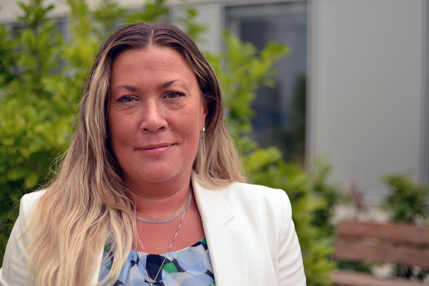 Leende kvinna i en blå blus och en vit kavaj ståendes ute i naturen.