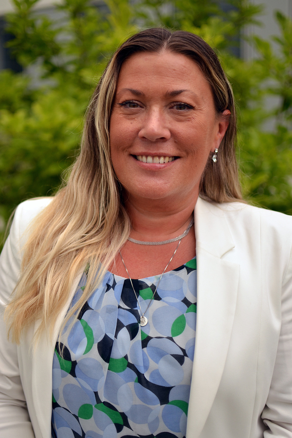 Leende kvinna i en blå blus och en vit kavaj ståendes ute i naturen.