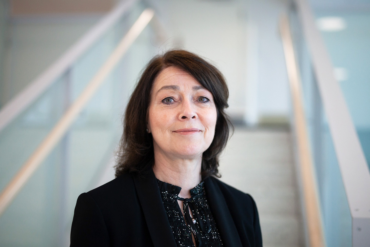 Kvinna i en glittrig blus med en svart kavaj över ståendes i ett fint ljust trapphus. 