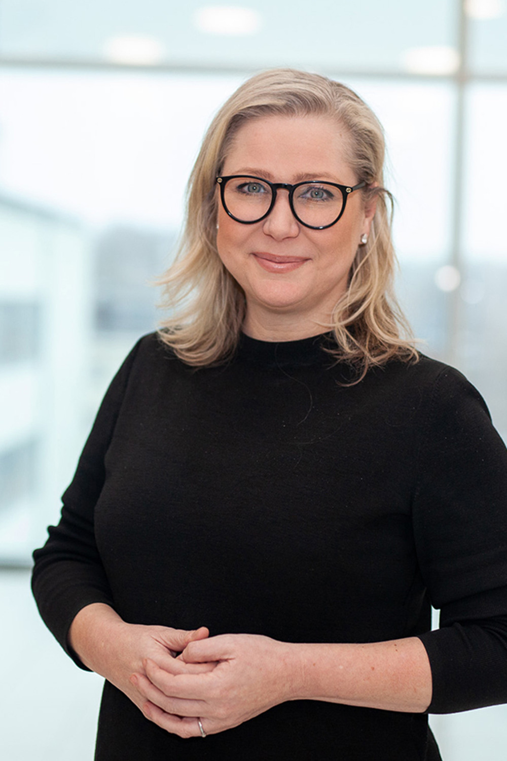 Kvinna med svarta glasögon och en svart halvpolo ståendes i ett fint ljust trapphus med knäppta händer.
