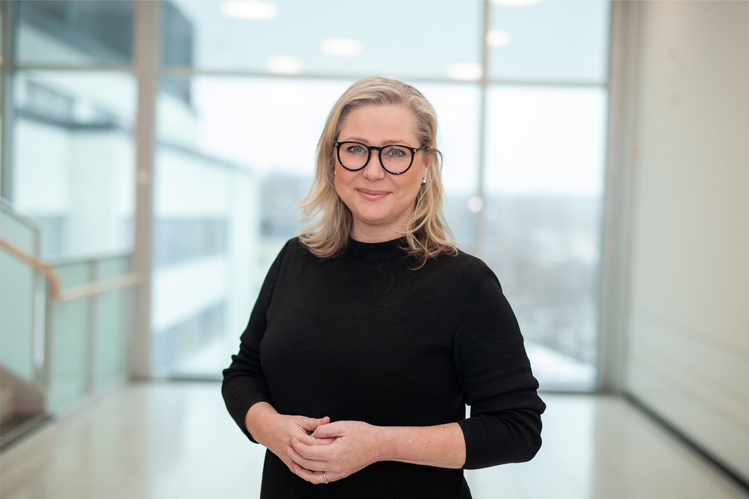 Kvinna med svarta glasögon och en svart halvpolo ståendes i ett fint ljust trapphus med knäppta händer.