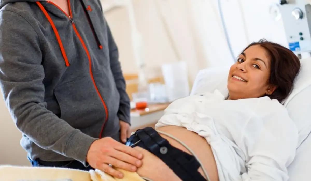 Gravidkvinna som ligger i sjukhussäng med en man bredvid som känner på magen. 
