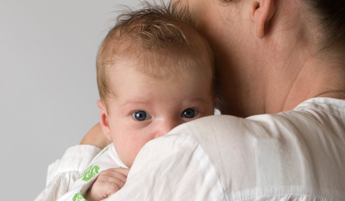 En bebis som kikar fram bakom en axel och kolla rakt in i kameran.