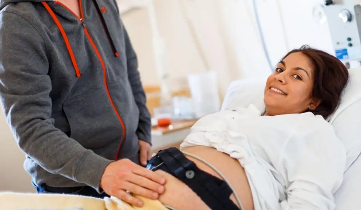 Gravidkvinna som ligger i sjukhussäng med en man bredvid som känner på magen. 