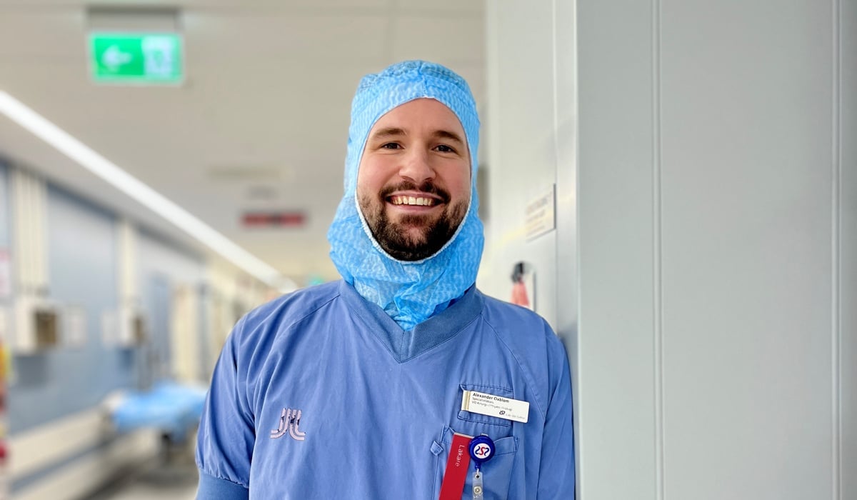 Glad man iklädd blåa vårdkläder och en blå operationsmössa ståendes i en korridor utanför operationssalarna.