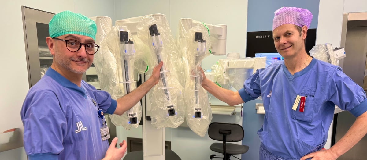 Två leende män i operationskläder står vid en operationsrobot