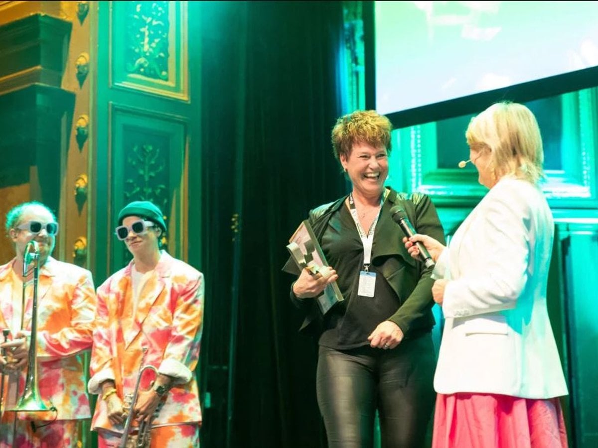 En glad kvinna som tar emot ett pris med två musiker bakom sig.